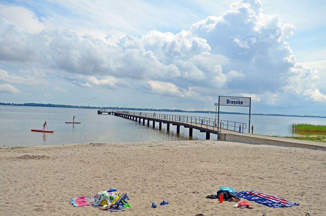 A beach with a pier extending into calm waters, a sign reading 'Dranske,' and beachgoers with beach gear.