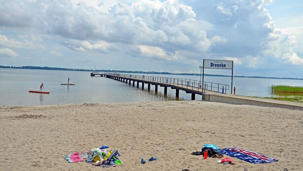 Ein Strand mit einem Steg, der sich über ruhiges Wasser erstreckt, einem Schild mit der Aufschrift 'Dranske' und Erholungssuchenden mit Strandausrüstung.