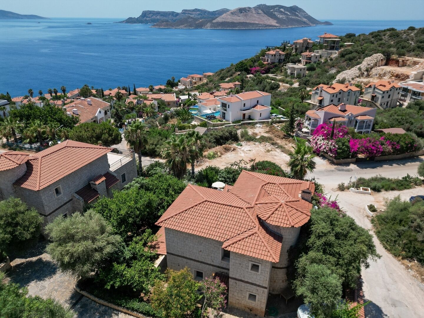 A coastal residential area with houses featuring red-tiled roofs, surrounded by greenery and a view of the sea.