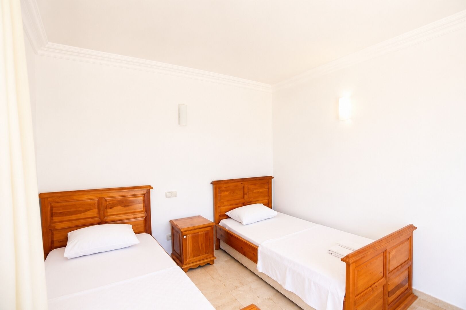A room with two single beds, wooden headboards, white bedding, a small wooden nightstand, and a light switch on the wall.