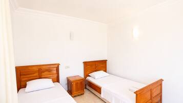 A room with two single beds, wooden headboards, white bedding, a small wooden nightstand, and a light switch on the wall.