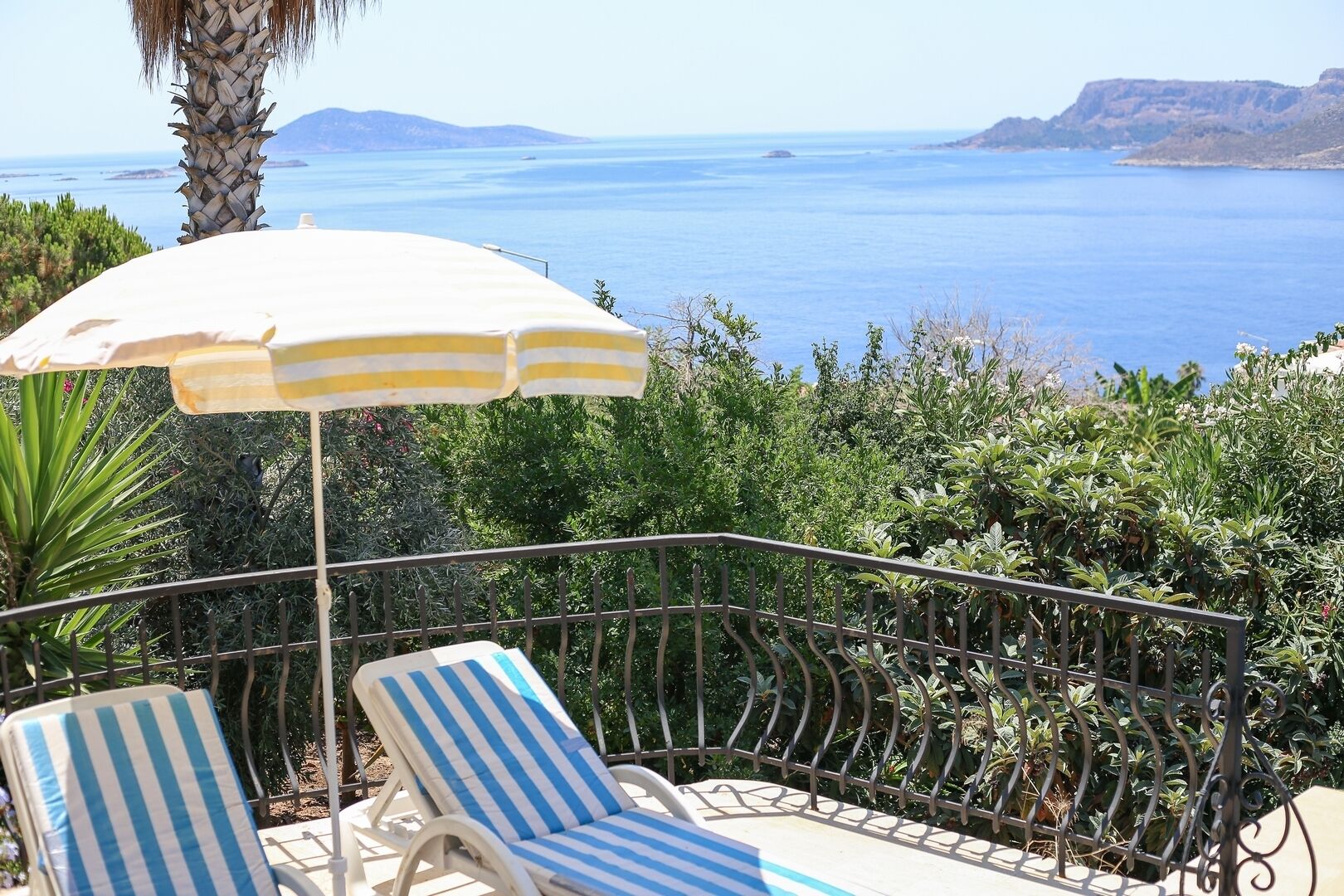 A balcony with two striped lounge chairs and a striped umbrella, overlooking a coastal landscape with clear blue sea and distant hills.