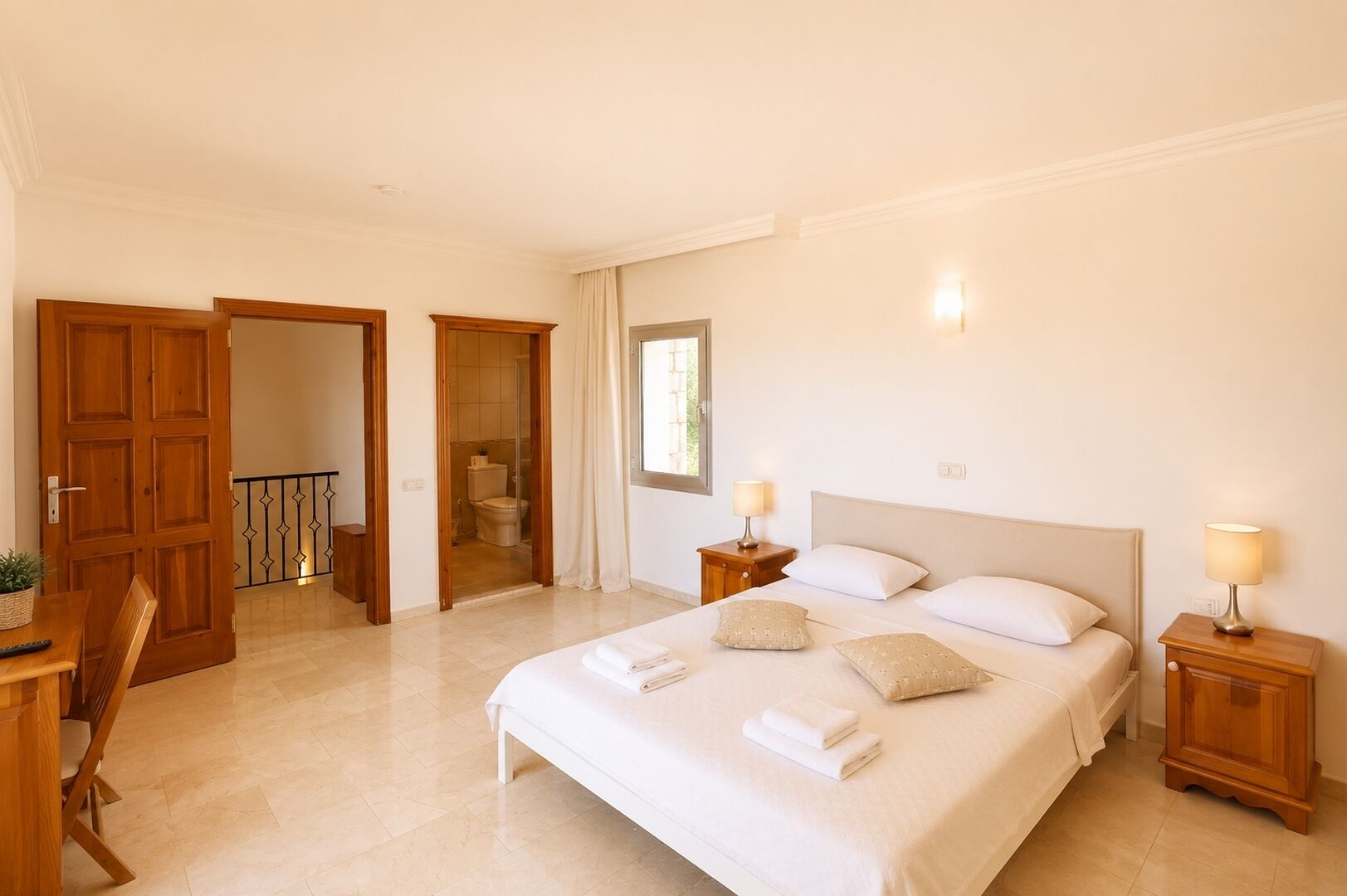Una habitación de hotel con una cama grande, mesitas de noche con lámparas, un baño visible a través de una puerta abierta y un área de comedor con mesa y sillas.