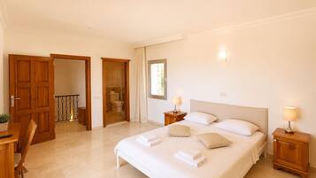 Una habitación de hotel con una cama grande, mesitas de noche con lámparas, un baño visible a través de una puerta abierta y un área de comedor con mesa y sillas.
