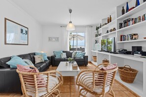 A modern living room with a black leather sofa, two wicker chairs, a white coffee table, and a bookshelf filled with books and decorative items. - Vacation Home 'Casa Canas' with Sea View, Wi-Fi and Air Conditioning (Playa Blanca)