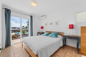 A bedroom with a bed, a desk, a chair, a window with curtains, and a view of a pool and palm trees. - Vacation Home 'Casa Canas' with Sea View, Wi-Fi and Air Conditioning (Playa Blanca)
