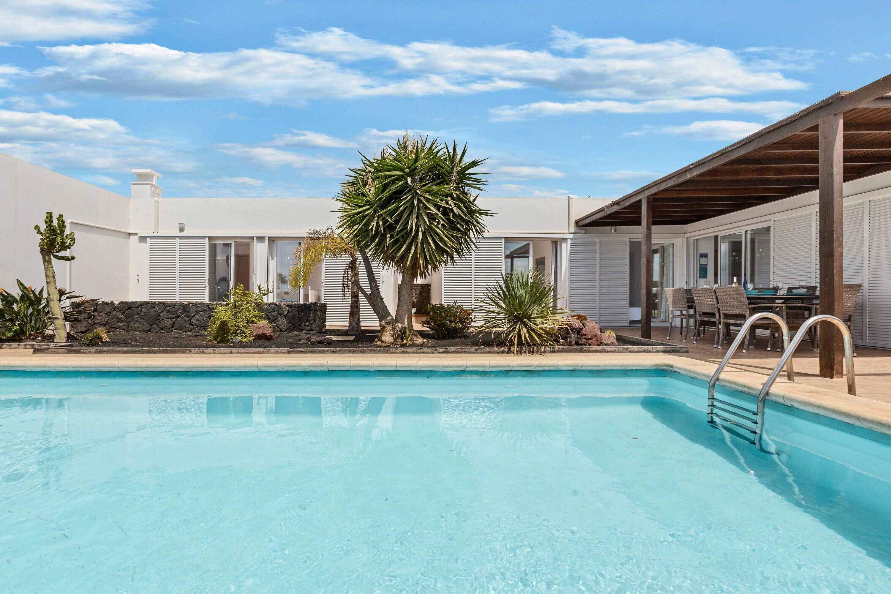 Une piscine dotée d’une terrasse en bois et de chaises longues, entourée de plantes tropicales et bordée d’un bâtiment blanc aux volets fermés.