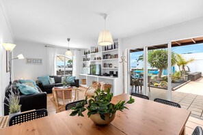 A modern living room with a wooden dining table, black sofa, and wicker chairs. A bookshelf filled with books and decorative items is visible, and a large window offers a view of the outdoors. - Vacation Home 'Casa Canas' with Sea View, Wi-Fi and Air Conditioning (Playa Blanca)