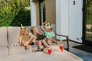Three children sitting on a beige sofa outdoors with a glass of red drink on a table in front of them.
