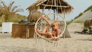 Iemand zit in een hangstoel op een zandstrand, met een rieten constructie op de achtergrond.