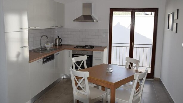 A compact kitchen with white cabinets, a gas stove, a sink, and a dining area with a wooden table and white chairs. - Apartments Vala (Grebaštica)