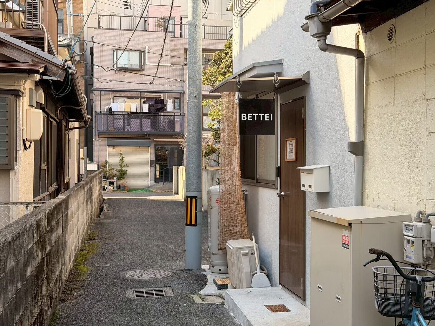 A narrow alleyway with a building labeled 'BETTEI' on the right, a bicycle parked outside, and a manhole cover on the ground.