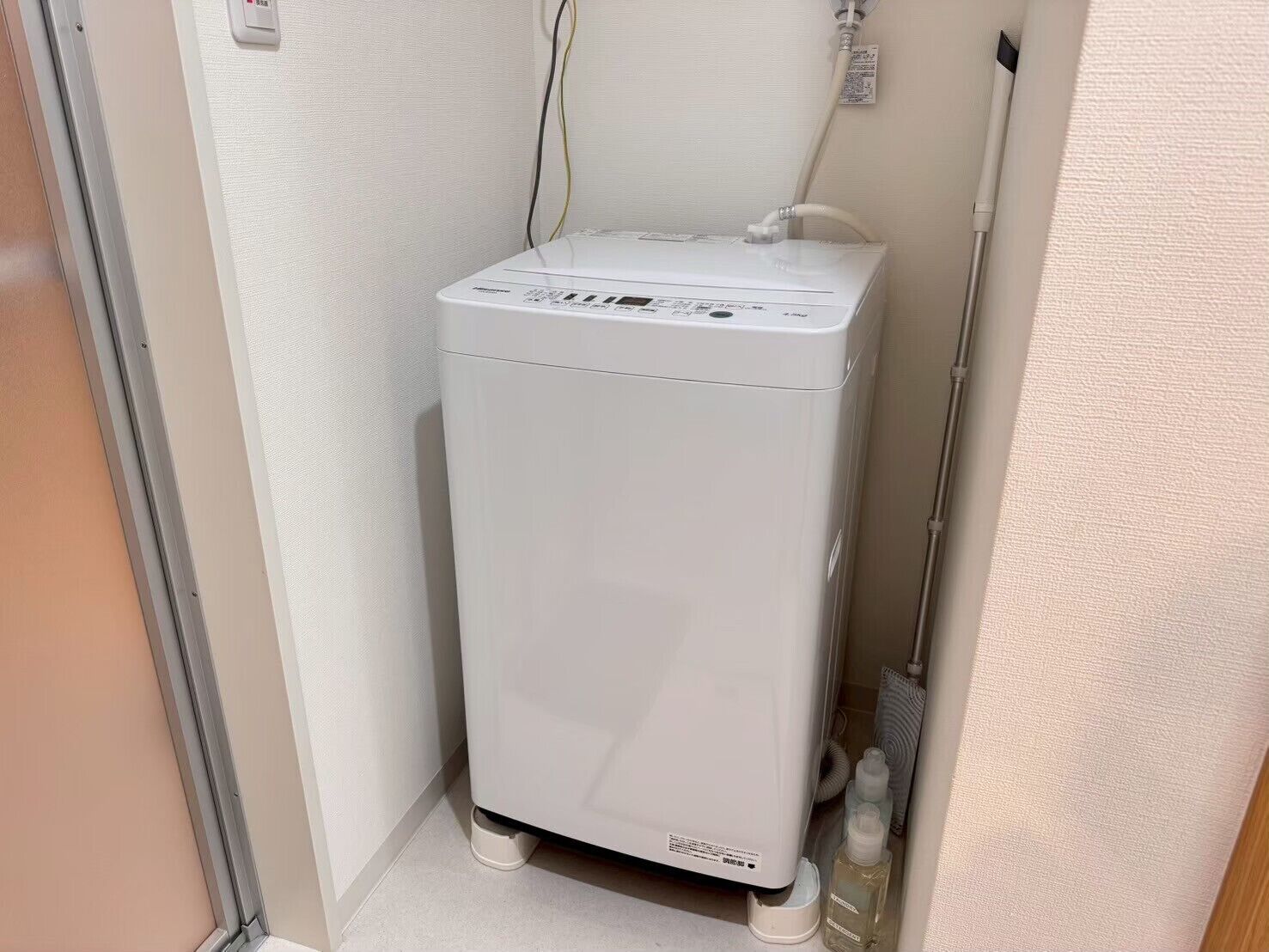 A compact, white, wall-mounted water heater in a corner of a room with a door and a towel rack.