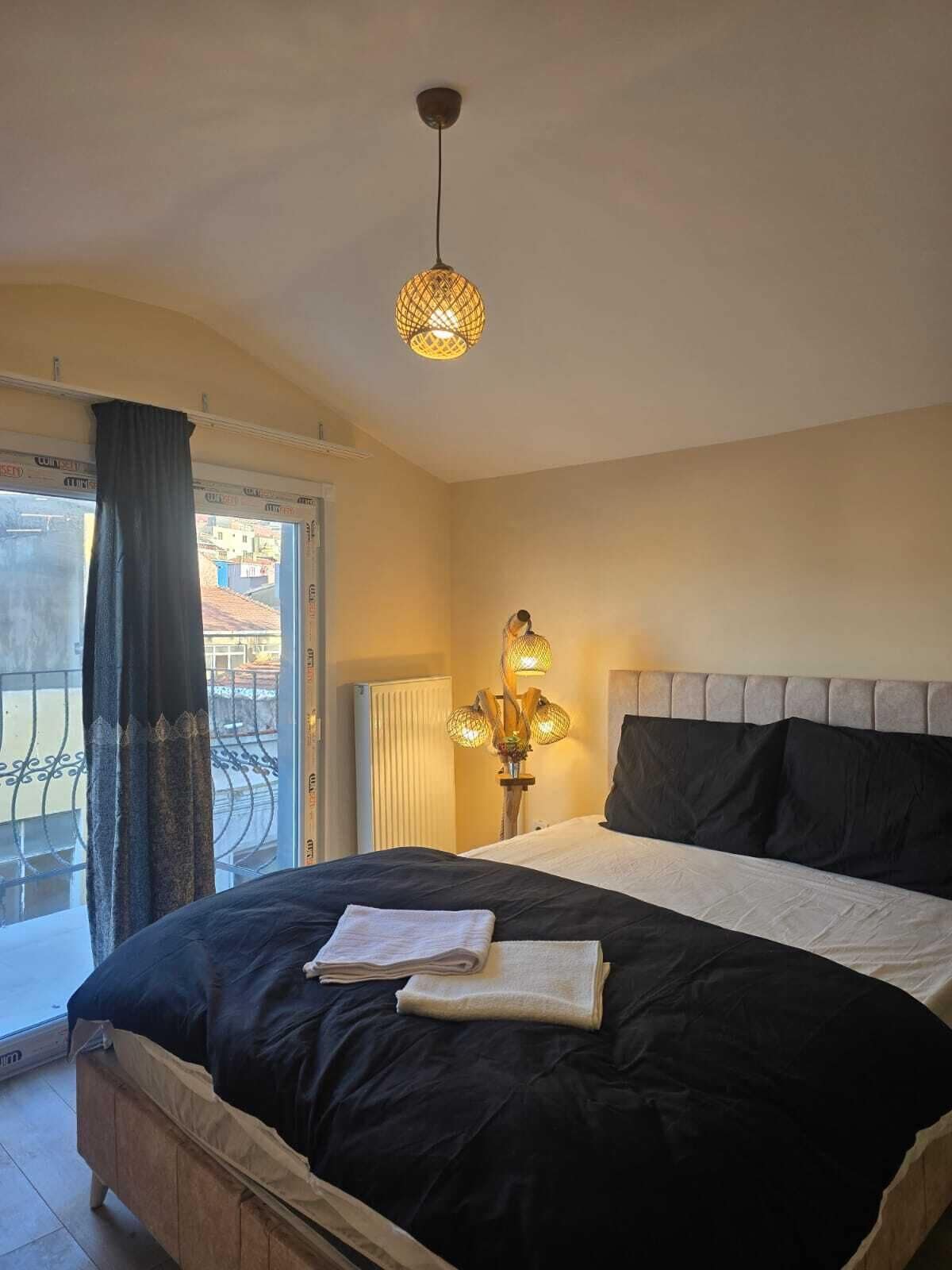 A hotel room with a bed, a window with curtains, a radiator, and a hanging light fixture.