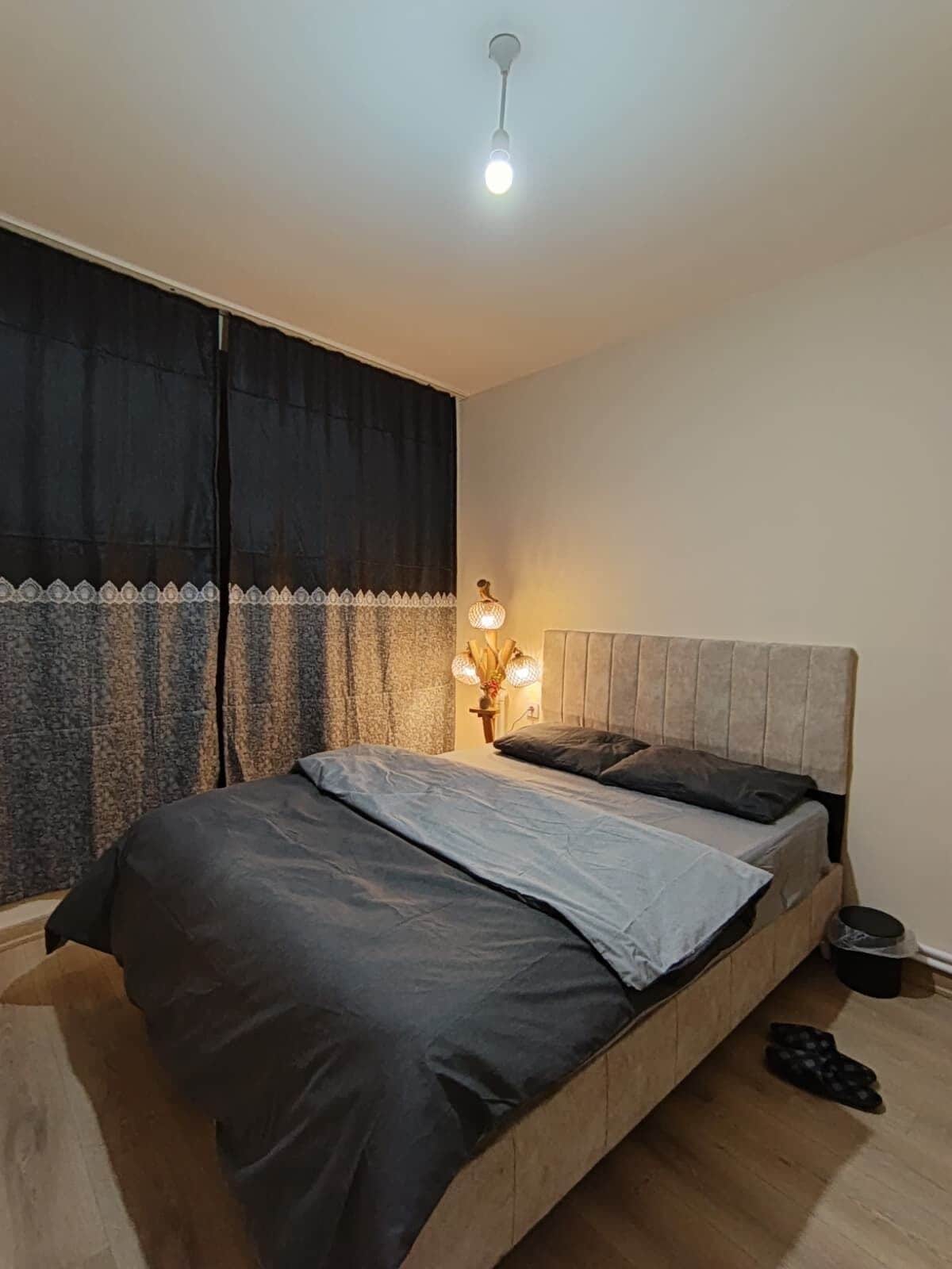 Un dormitorio con cama, mesita de noche, lámpara y ventana con cortinas.