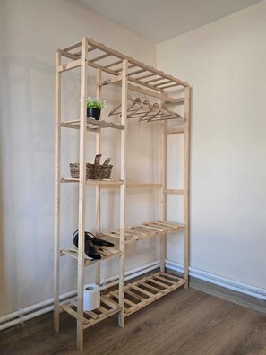 A wooden shelving unit with multiple shelves and a hanging rack, positioned against a plain wall with a wooden floor.