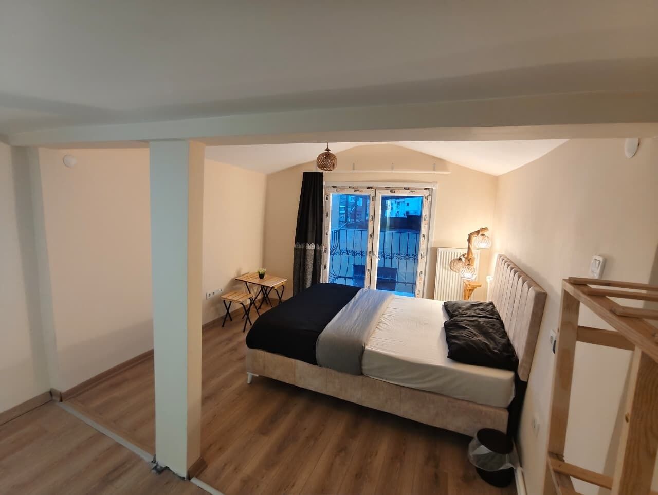 A bedroom with a bed, a small table, a chair, a radiator, and a window with a view.