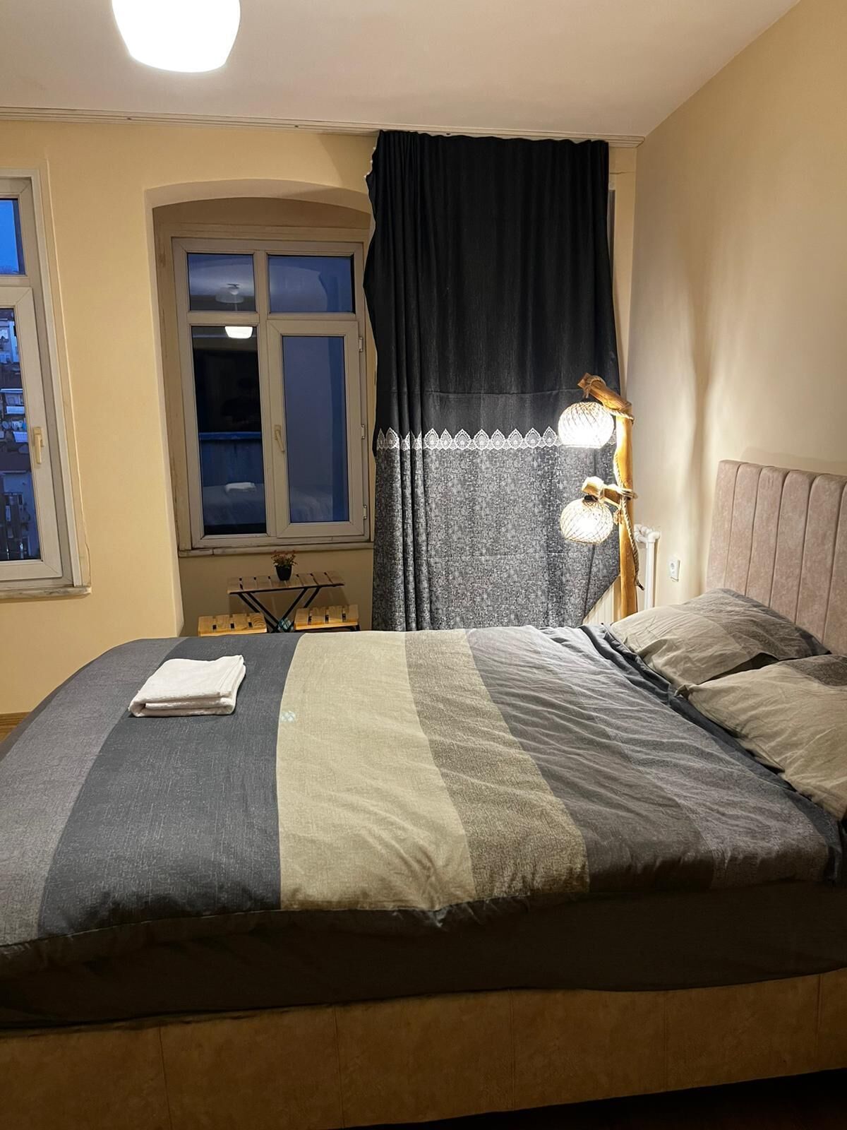 A bed with a blanket, a bedside lamp, a window with curtains, and a nightstand with a lamp.