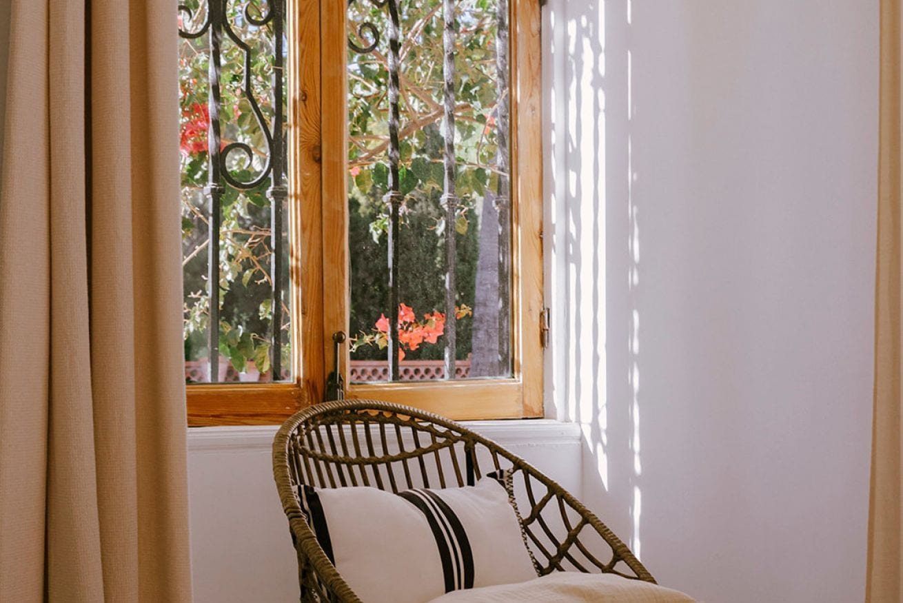 Une chaise en osier avec un coussin, placée devant une fenêtre donnant sur des arbres et des fleurs.