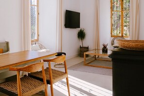 A room with a wooden dining table, chairs, a television mounted on the wall, a potted plant, and a window with curtains. - Hacienda Clavero: A Garden Apartment (Málaga)