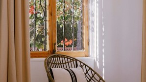 Una silla de mimbre con cojín frente a una ventana con vista a árboles y flores.