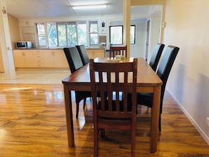 A dining area with a wooden table and chairs, a kitchen in the background, and a view of the outdoors through the windows.