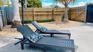 Two lounge chairs with cushions, a wooden fence, a tree, and a paved patio area.