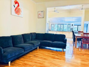 A living room with a dark-colored sectional sofa, wooden flooring, and a dining area with a table and chairs. - 2- bedroom house in tranquil Rockhampton with Pool (Rockhampton)