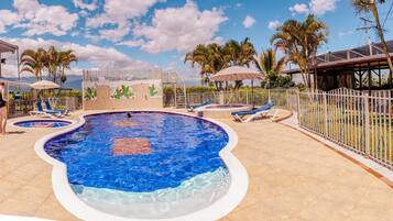 A swimming pool with lounge chairs and umbrellas, surrounded by palm trees and a white fence.
