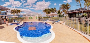 A swimming pool with lounge chairs and umbrellas, surrounded by palm trees and a white fence. - Meraki is a Luxury Villa with pool, jacuzzi & Stunning Mountain Views (Quindío)
