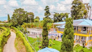 Un edificio de dos pisos con techo azul, rodeado de vegetación y un camino de tierra.