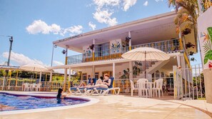 A pool area with lounge chairs and umbrellas, a covered seating area, and a building with a balcony.