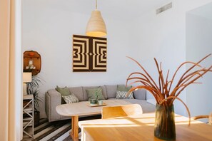 A modern living room with a sofa, a wooden table, a hanging light fixture, and a decorative wall piece. - Hacienda Clavero Two Garden Apartment (Málaga)