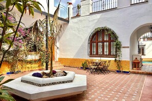 A courtyard with a stone planter, outdoor seating, and a building with a white facade and arched window. - Hacienda Clavero Three Patio Apartment: (Málaga)