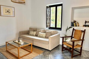 A living room with a sofa, a wooden chair, a glass coffee table, a window with a view, and a bookshelf with books. - Hacienda Clavero Three Patio Apartment: (Málaga)
