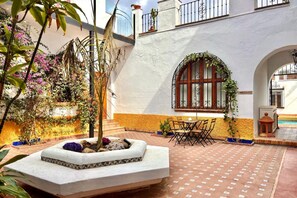 A courtyard with a stone planter, outdoor seating, and a building with a white facade and arched window. - Hacienda Clavero Three Patio Apartment: (Málaga)