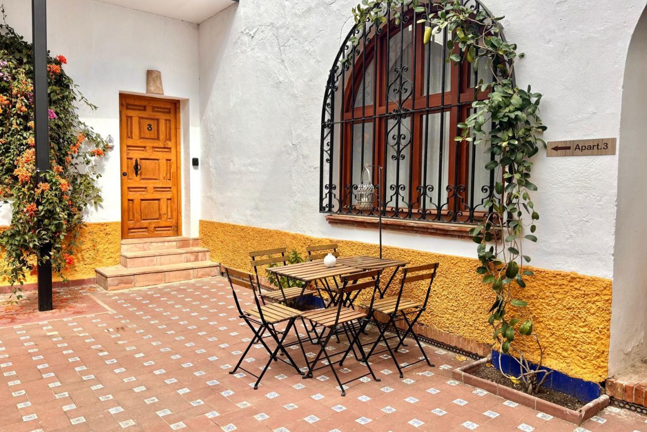 Ein Innenhof mit einer Holztür, einem Fenster mit Eisengittern und einem Tisch mit Stühlen.