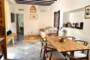 A modern living room with a wooden dining table, cushioned seating, a hanging light fixture, and a bookshelf with books and decorative items. - Hacienda Clavero Three Patio Apartment: (Málaga)
