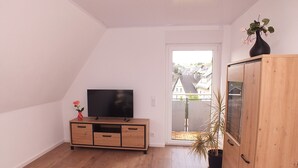 Habitación con televisor sobre un mueble de madera, una planta en maceta y vistas a través de un ventanal.