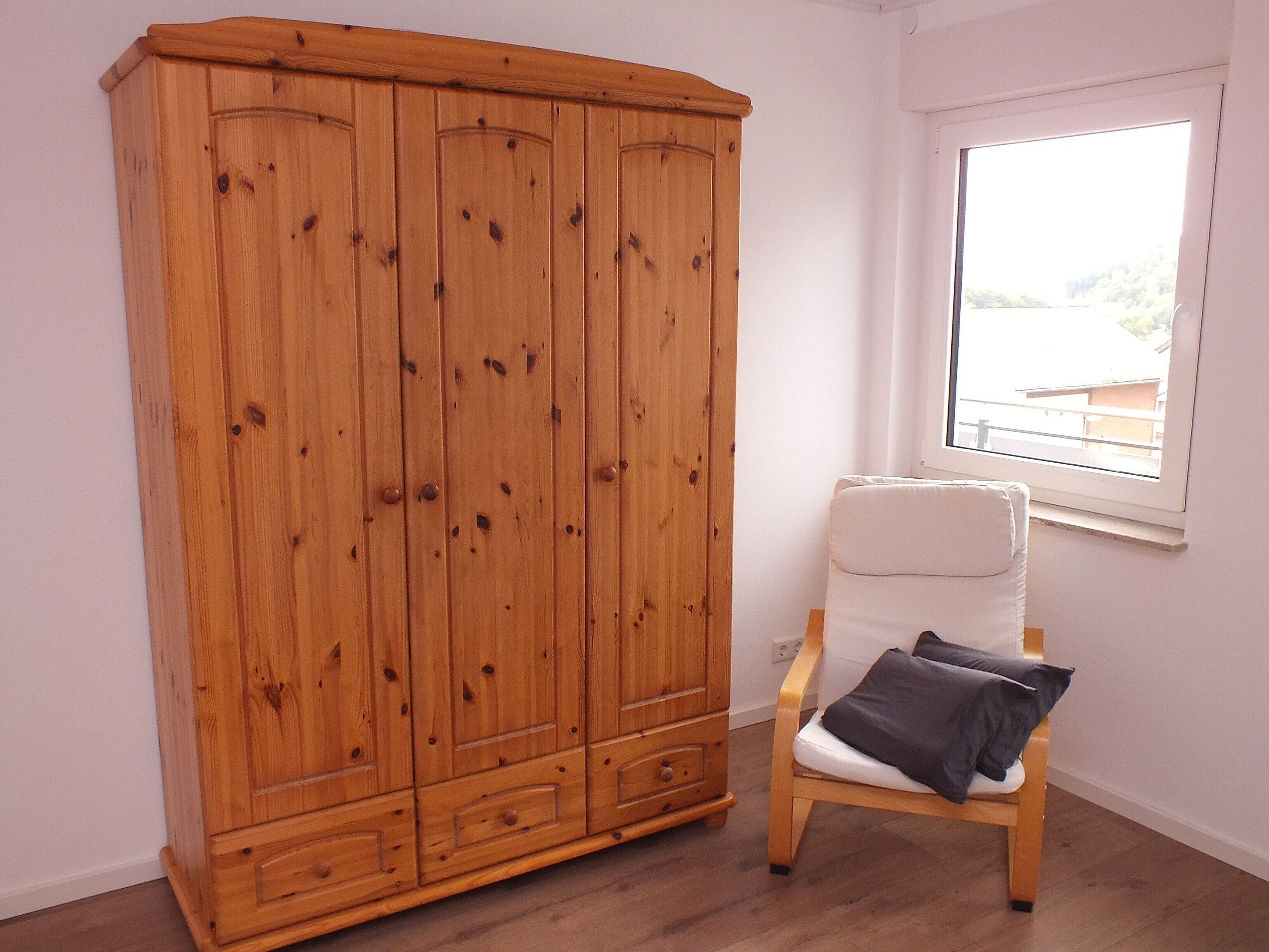 Une pièce avec une armoire en bois, une chaise blanche avec un coussin et une fenêtre donnant sur un beau paysage.