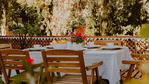 Un espacio de comedor al aire libre con sillas de madera y una mesa cubierta con mantel blanco, platos y un centro de mesa con flores.