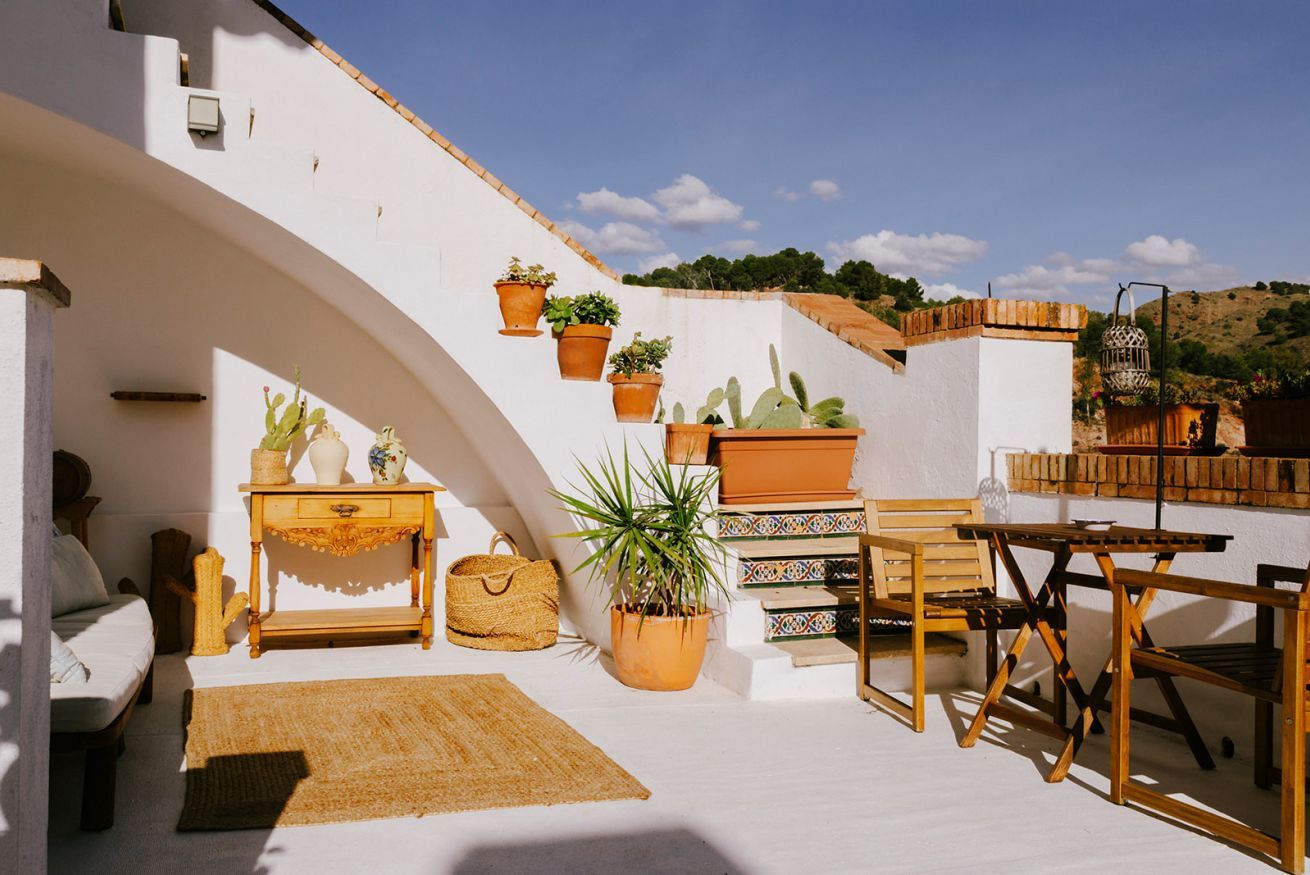 Eine sonnendurchflutete Terrasse mit weißer Wand, Holzmöbeln und Pflanzen in Töpfen.