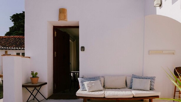 Pared exterior blanca con un banco de madera, una mesa pequeña con planta en maceta y un acceso a un espacio interior.