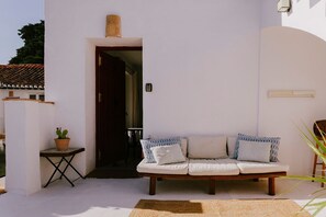 Paredes externas brancas com um banco de madeira, uma mesa pequena com um vaso de planta e uma porta de entrada para um espaço interno.