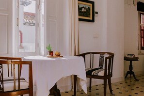Un comedor con una mesa dispuesta para dos, una silla, una mesita con una planta y un cuadro en la pared.