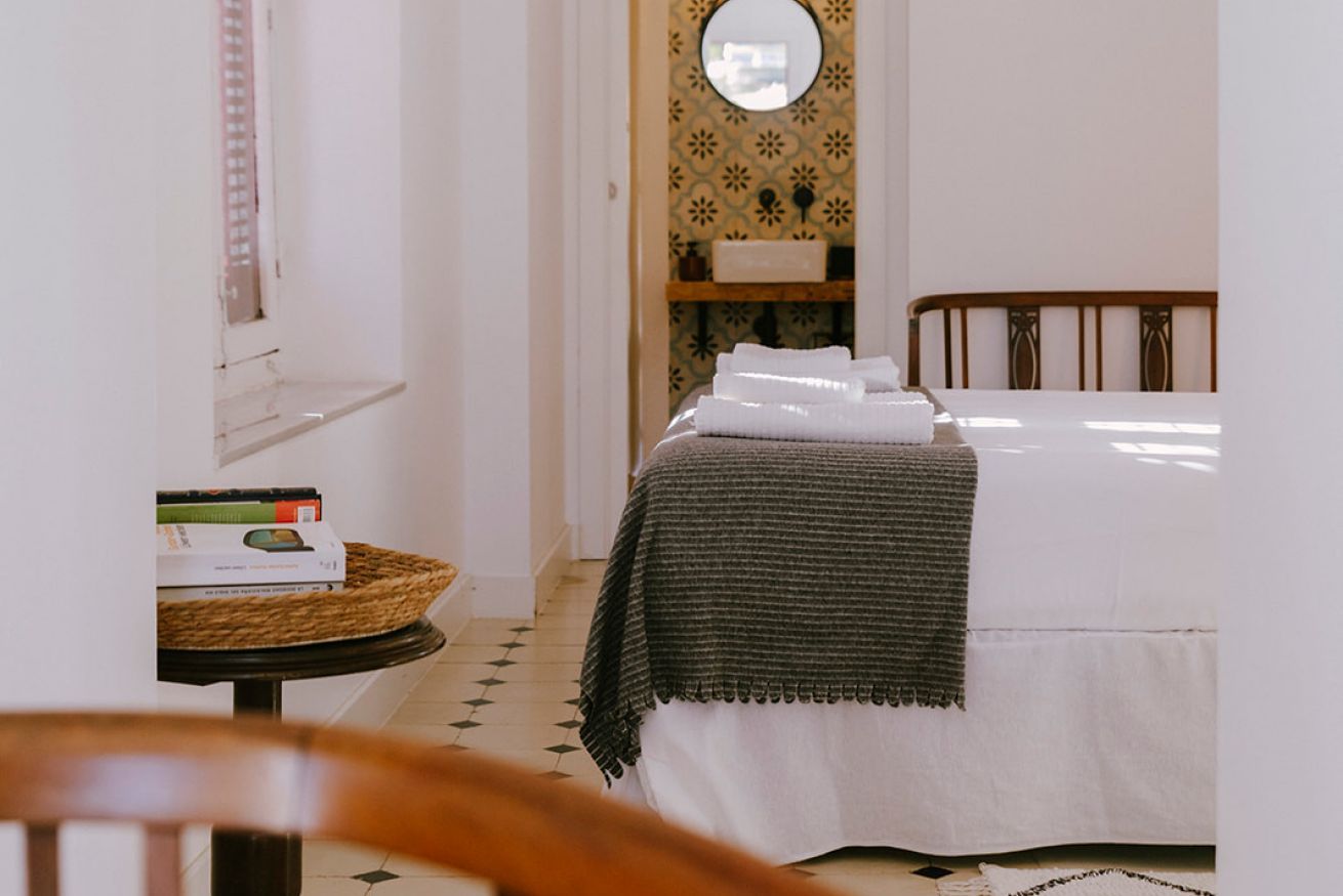 Ein ordentlich bezogenes Bett mit weißer und dunkelgrüner Decke, ein Nachttisch mit Büchern und einem gewebten Korb, sowie eine Wand mit Muster und einem Spiegel über dem Bett.