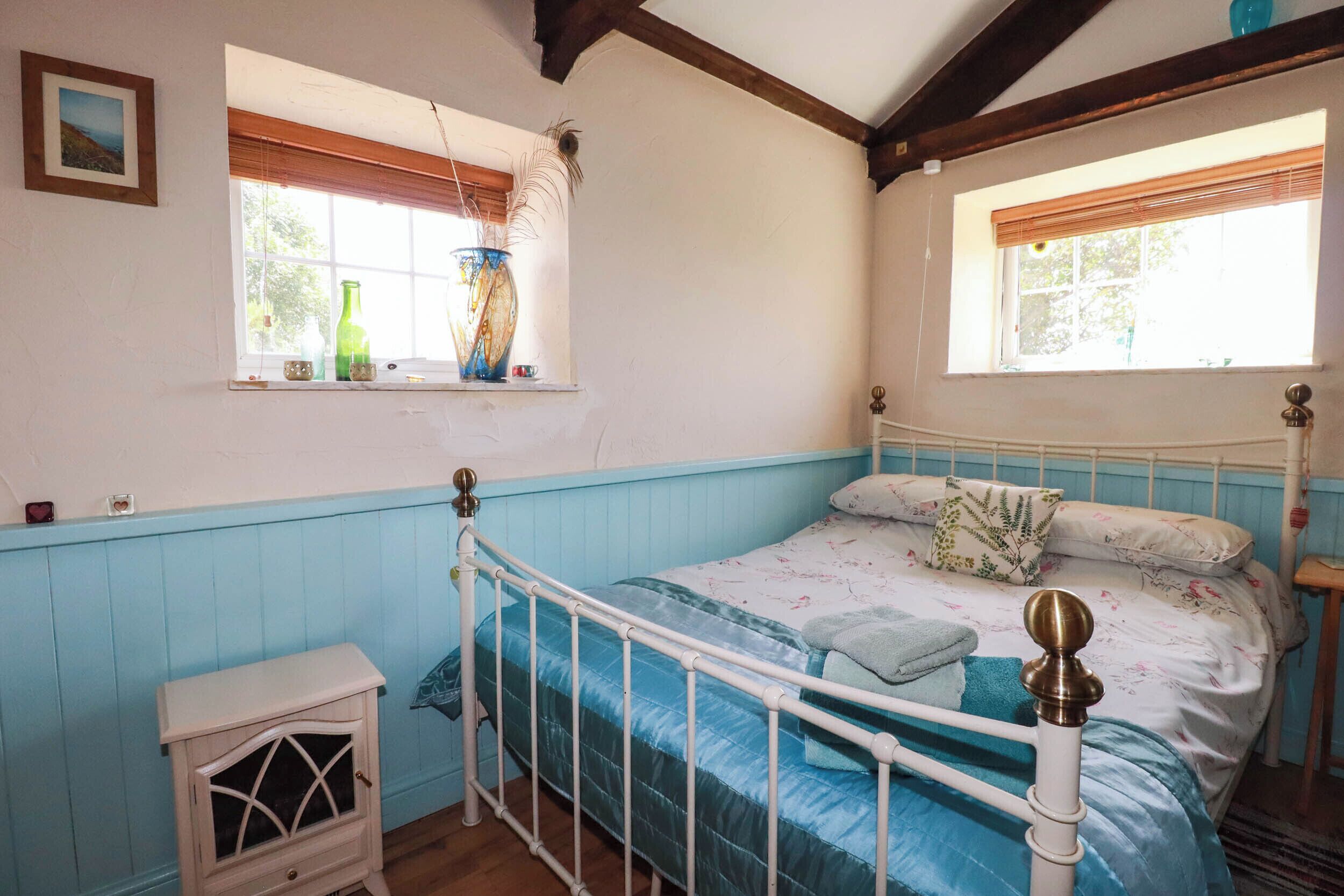 Un dormitorio con cama, mesita de noche, ventana con cortinas e imagen en la pared.