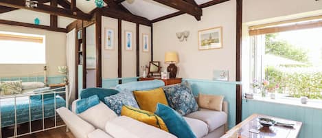 A room with a wooden ceiling, a bed with a blue bedspread, a sofa with multiple cushions, a small table with a lamp, and a window with a view of greenery.