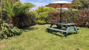 Una mesa de picnic en un área verde con un paraguas grande y diversas plantas y arbustos al fondo.