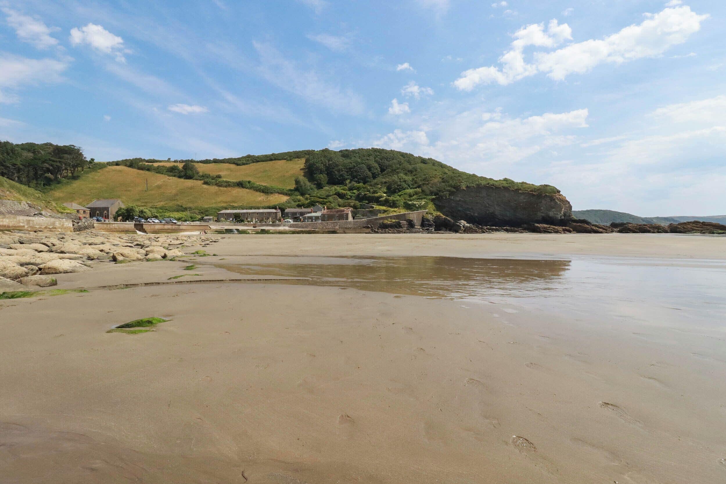 A sandy beach with a rocky shoreline, a small building, and a hill with vegetation in the background.
